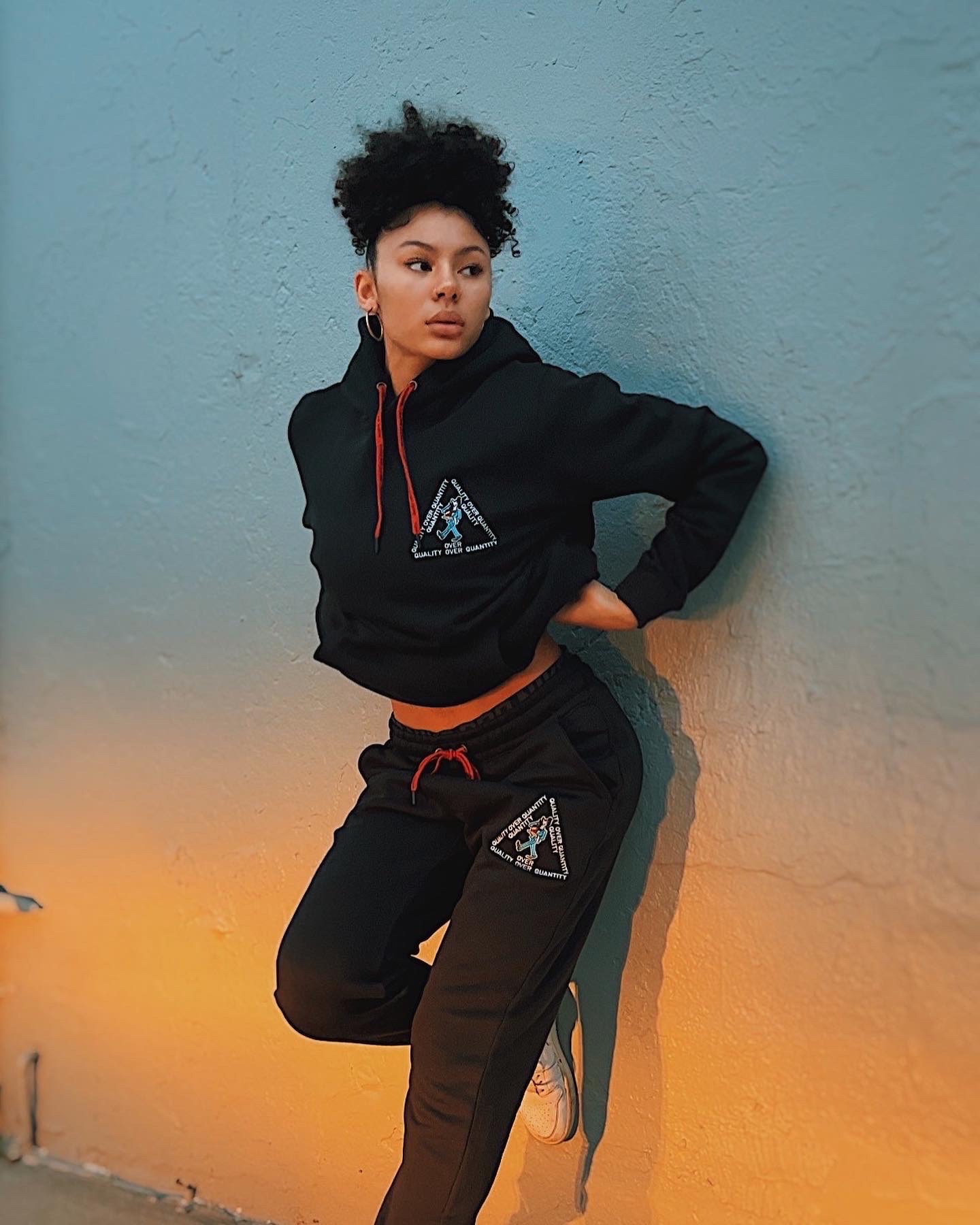 Model leaning against painted wall in PETRO QOQ hoodie and joggers, lit by warm sunset